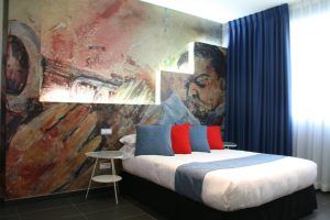 El Hotel Indiana de Madrid presenta una cautivadora habitación con un vibrante mural de temática jazz, con una cama adornada con almohadas azules y rojas y elegantes cortinas azules. - ForTwo - Planes para parejas