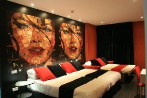 Descubra la elegancia moderna del Hotel Indiana en Madrid. Nuestra elegante habitación con dos camas individuales presenta una impactante decoración en rojo y negro, acentuada con cuadros de rostros femeninos abstractos. - ForTwo - Planes para parejas