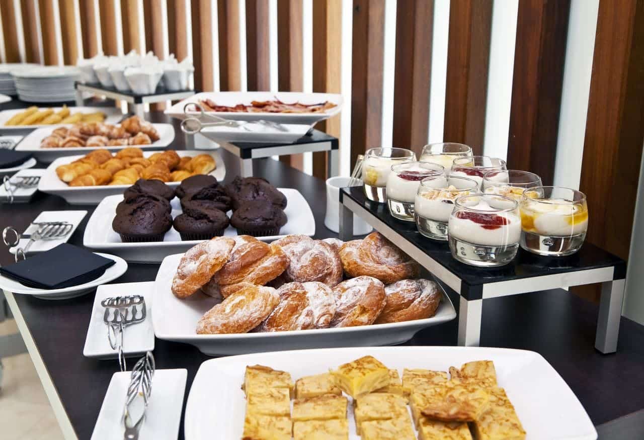 El buffet del Hotel Meliá Sol y Nieve en Sierra Nevada (Granada) cuenta con una selección de pasteles, magdalenas y vasitos de postre elegantemente presentados en platos y bandejas blancas. - ForTwo - Planes para parejas
