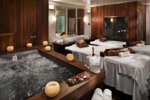 Experimente la máxima relajación en el Hotel Meliá Sol y Nieve en Sierra Nevada, con una lujosa sala de spa con bañera de hidromasaje, mesas de masaje, champán y una bandeja de frutas en un mostrador de madera, todo ello realzado por el relajante resplandor de las velas. - ForTwo - Planes para parejas