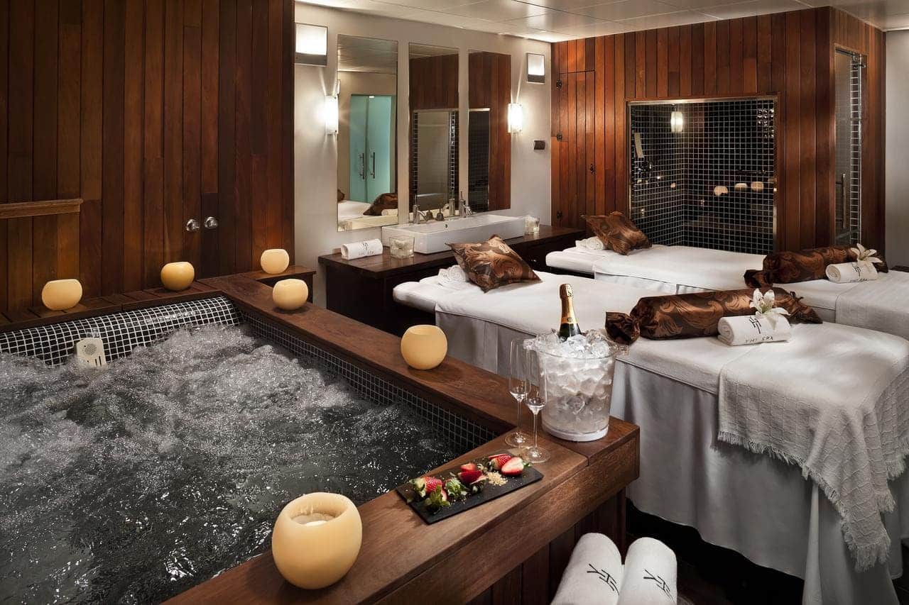 Experimente la máxima relajación en el Hotel Meliá Sol y Nieve en Sierra Nevada, con una lujosa sala de spa con bañera de hidromasaje, mesas de masaje, champán y una bandeja de frutas en un mostrador de madera, todo ello realzado por el relajante resplandor de las velas. - ForTwo - Planes para parejas