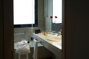 Este baño, que recuerda al Hotel Paseo del Arte de Madrid, exhibe una toalla en un taburete, artículos de tocador en el lavabo y flores de color rojo vivo elegantemente colocadas en un jarrón junto a la ventana. - ForTwo - Planes para parejas