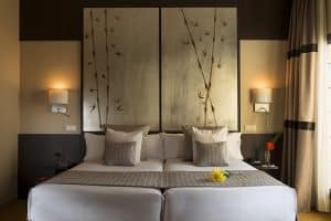 Disfrute de una habitación moderna en el Hotel Paseo Del Arte de Madrid, equipada con una cama doble, una decoración elegante, arte en bambú, una iluminación suave y una acogedora flor amarilla en la cama. - ForTwo - Planes para parejas