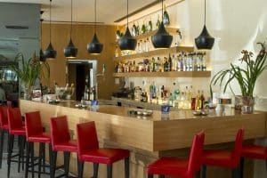 Un moderno bar en el Hotel Paseo Del Arte (Madrid) cuenta con un mostrador de madera, sillas rojas, lámparas colgantes negras y estanterías llenas de botellas. - ForTwo - Planes para parejas