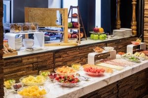 En el Hotel Meliá Princesa Madrid, un delicioso buffet en una encimera de mármol incluye frutas en rodajas, quesos y pasteles, complementados con una exhibición de frutas y platos apilados en el fondo. - ForTwo - Planes para parejas