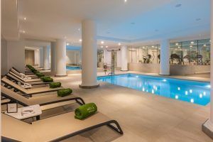 El Hotel Meliá Princesa de Madrid ofrece una piscina cubierta con tumbonas adornadas con toallas de color verde vibrante, una vista de fondo de un gimnasio con paredes de cristal y una iluminación suave que realza el ambiente sereno, creando un oasis lejos del bullicio de la ciudad. - ForTwo - Planes para parejas