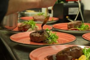 En el Restaurante Alacena de las Monjas de Granada, un chef vierte elegantemente salsa sobre un filete en un plato rojo con verduras, mostrando el famoso Menú Aljibe. - ForTwo - Planes para parejas