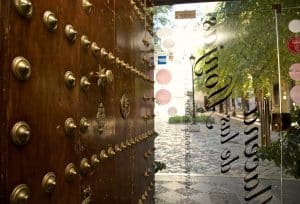 Una puerta de madera ornamentada se abre a una soleada calle adoquinada de Granada, bordeada de árboles y tiendas con encanto, que invita a explorar joyas cercanas como el Menú Aljibe del Restaurante Alacena de las Monjas. - ForTwo - Planes para parejas