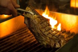 Una mano agarra un filete a la parrilla con unas pinzas sobre una llama abierta, evocando los sabores ardientes del Menú Aljibe en el Restaurante Alacena de las Monjas en Granada. - ForTwo - Planes para parejas