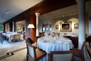 El Restaurante Abades en Ronda ofrece una experiencia de menú romántico con elegantes interiores que presentan mesas redondas con manteles blancos, equipadas con cristalería y platos, complementados con arte en las paredes. - ForTwo - Planes para parejas