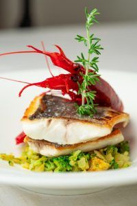 Saborea el Menú romántico en el restaurante Abades (Ronda), que incluye un filete de pescado a la parrilla gourmet con una gamba grande y brócoli, elegantemente adornado con romero en un plato blanco, perfecto para abrazar el ambiente romántico. - ForTwo - Planes para parejas