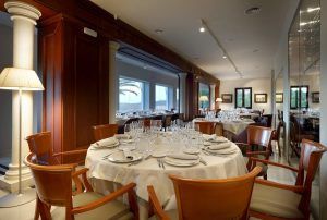 El elegante interior del Restaurante Abades cuenta con una mesa redonda con mantel blanco y sillas de madera, perfecta para el Menú romántico del restaurante Abades (Ronda). Los amplios ventanales que se encuentran tras ella ofrecen una vista cautivadora del encantador paisaje de Ronda. - ForTwo - Planes para parejas