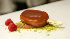 En el Restaurante Azahar de Sevilla, saboree una barra de helado de caramelo sobre crumble de pistacho con frambuesas frescas, bellamente presentada en un impecable plato blanco. - ForTwo - Planes para parejas