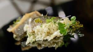En el Restaurante Azahar de Sevilla, disfrute de una comida gourmet con un tierno filete de pescado, un risotto cremoso, hierbas frescas y esferas verdes decorativas bellamente servidas en un elegante plato negro. - ForTwo - Planes para parejas