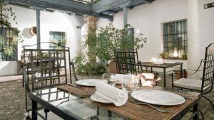 Encantadora cafetería con patio, con mesas de metal para dos, rodeada de exuberante vegetación y arquitectura colonial, que evoca la elegancia sevillana. Esta joya escondida encarna el espíritu del Restaurante Azahar en cada detalle. - ForTwo - Planes para parejas