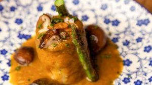 Plato gourmet con salsa, espárragos, champiñones y hierbas elegantemente dispuestos en un plato decorativo azul y blanco en el Restaurante Bache San Pedro (Sevilla). - ForTwo - Planes para parejas