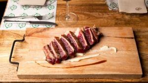 En Restaurante Bache San Pedro (Sevilla), disfrute de un filete cortado en rodajas servido en una tabla de madera con salsa, presentado en una mesa de madera con cubiertos y una copa de vino cerca. - ForTwo - Planes para parejas