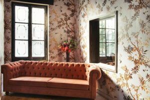 Una habitación del Restaurante Küche de Madrid incluye un sofá naranja con mechones acolchados, papel tapiz con motivos florales, un gran espejo y una ventana con un ramo de flores al lado. - ForTwo - Planes para parejas