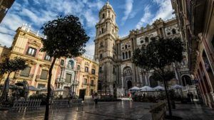 Descubra el encanto de una histórica plaza europea en Málaga, donde los árboles dan sombra a las terrazas y la imponente catedral se yergue imponente. Disfrute de una cena en el Restaurante L'Expérience mientras se sumerge en el vibrante ambiente bajo un cielo azul con nubes. - ForTwo - Planes para parejas