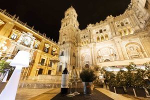 Una impresionante vista nocturna de un edificio histórico iluminado de Málaga sirve de telón de fondo para Restaurante L'expérience, invitándole a saborear un buen vino en una mesa bellamente dispuesta en primer plano. - ForTwo - Planes para parejas