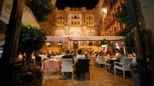 Los clientes disfrutan cenando en el Restaurante L'expérience en Málaga, disfrutando de las vistas nocturnas de una catedral histórica iluminada mientras están sentados bajo un toldo rodeado de mesas y plantas, creando un ambiente encantador. - ForTwo - Planes para parejas