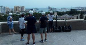 La gente en una cornisa contempla el paisaje urbano y el puerto de Málaga, con cascos y de pie junto a vehículos estacionados, listos para la emocionante Ruta en segway por Málaga. - ForTwo - Planes para parejas