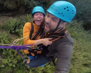Dos personas, ataviadas con casco y riendo alegremente, se embarcan en un emocionante "Salto puenting tandem en pareja en Saldes" entre una exuberante vegetación. - ForTwo - Planes para parejas