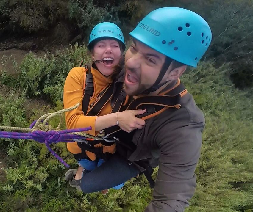 Dos personas, ataviadas con casco y riendo alegremente, se embarcan en un emocionante "Salto puenting tandem en pareja en Saldes" entre una exuberante vegetación. - ForTwo - Planes para parejas
