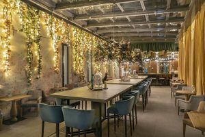 Disfrute de una acogedora experiencia gastronómica en el interior del Hotel Vincci Soho de Madrid. El espacio está decorado con flores y luces, con largas mesas y sillas bajo un techo transparente para una comida íntima. - ForTwo - Planes para parejas