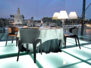 Disfrute de una cena romántica de lujo en el Restaurante Abades Triana (Sevilla), que cuenta con un elegante comedor y suelo de cristal con impresionantes vistas al río y a la torre histórica al anochecer. - ForTwo - Planes para parejas
