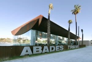 Un edificio moderno con fachada de cristal, palmeras y un destacado cartel de Restaurante Abades Triana en frente, perfecto para disfrutar de una Cena de lujo romántica en el Restaurante Abades Triana (Sevilla) en el corazón de la ciudad. - ForTwo - Planes para parejas
