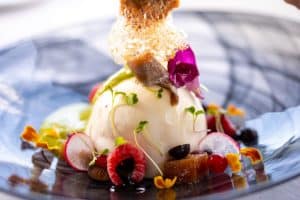 Disfrute de una elegante cena de lujo romántica en el Restaurante Abades Triana en Sevilla, donde una esfera cremosa está bellamente adornada con microvegetales, bayas, rodajas de rábano y pétalos de flores en un plato estampado mientras contempla las vistas panorámicas. - ForTwo - Planes para parejas