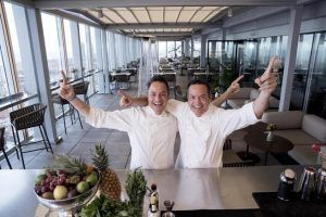 Dos cocineros del Restaurante Dos Cielos (Madrid) sonríen y señalan hacia arriba en una azotea, con un bol de fruta en primer plano. - ForTwo - Planes para parejas