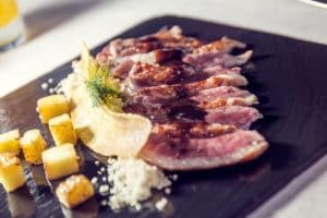 Disfrute de la Cena de lujo romántica en el Restaurante Abades Triana, Sevilla, con rodajas de carne a la parrilla en salsa en un plato negro, adornadas con hierbas, patatas cortadas en cubitos y un elegante remolino de crema. - ForTwo - Planes para parejas