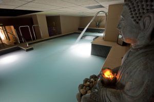 El Circuito Termal 90 minutos en Kumaras Spa (Umbrete) incluye una zona de piscina cubierta tenuemente iluminada con una serena cascada, escalones hacia el agua y una estatua de Buda sosteniendo una vela brillante, lo que mejora la experiencia de relajación. - ForTwo - Planes para parejas
