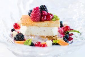 Una rebanada de tarta de queso adornada con frutos rojos reposa elegantemente sobre un plato de cristal, rodeada de frutas variadas y hojas de menta, creando el ambiente perfecto para una "Cena de lujo romántica en el Restaurante Abades Triana (Sevilla). - ForTwo - Planes para parejas