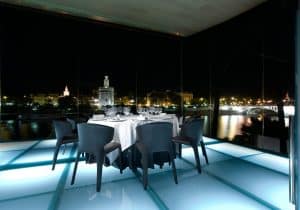 La lujosa Cena de lujo romántica en el Restaurante Abades Triana (Sevilla) ofrece un elegante comedor con una impresionante vista nocturna desde una plataforma de vidrio con vista al paisaje urbano y al río. - ForTwo - Planes para parejas