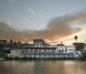 Una impresionante puesta de sol proyecta una cálida luz sobre la ciudad ribereña, fusionando la arquitectura tradicional sevillana con el encanto moderno. Disfrute de una "Cena de lujo romántica en el Restaurante Abades Triana" mientras el cielo vibrante se refleja en la serenidad del río, creando una velada inolvidable. - ForTwo - Planes para parejas