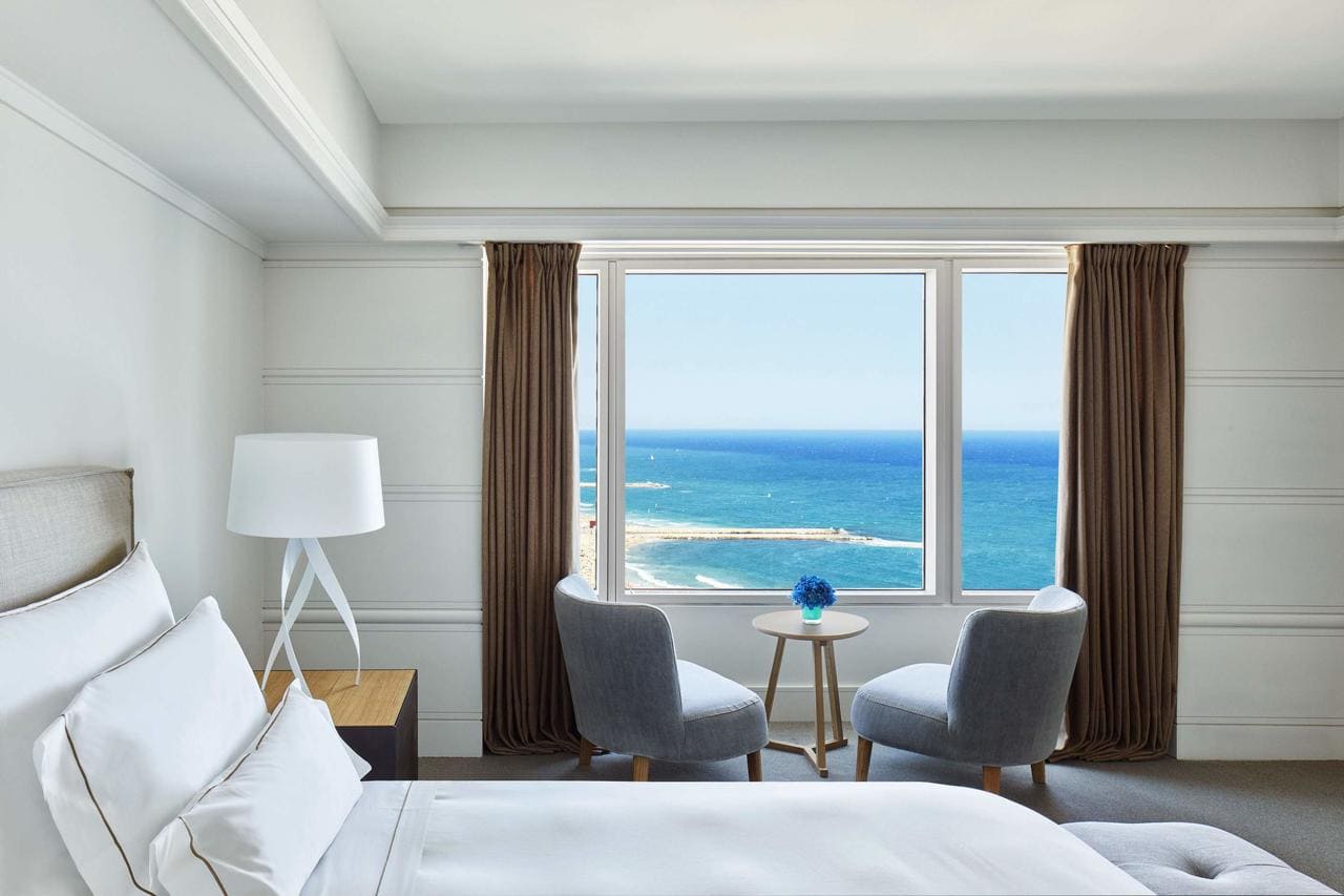 Una encantadora habitación costera en el Hotel Arts, Barcelona, incluye dos sillas junto a una ventana con vista al mar y una pequeña mesa adornada con un jarrón de flores azules. - ForTwo - Planes para parejas