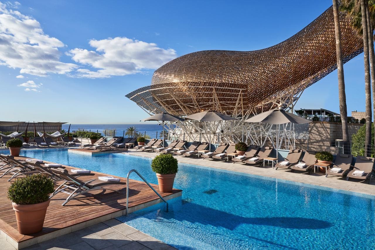 Disfrute de la moderna piscina del Hotel Arts Barcelona con tumbonas, sombrillas y una distintiva escultura en forma de pez bajo el impresionante cielo azul. - ForTwo - Planes para parejas