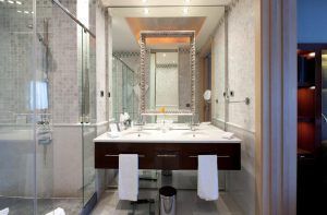 Recrea un baño inspirado en el Hotel Casa Fuster GL Monumento de Barcelona, con un lavabo doble, un espejo grande y una ducha de cristal. Las toallas cuelgan elegantemente sobre el lavabo bajo una suave iluminación ambiental. - ForTwo - Planes para parejas