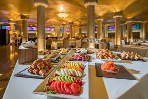Disfrute de un lujoso buffet en el Hotel Casa Fuster GL Monumento (Barcelona) con una variedad de frutas, pasteles y carnes en un elegante comedor realzado por majestuosas columnas. - ForTwo - Planes para parejas