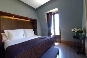 Relájese en una acogedora habitación del Hotel Casa Fuster GL Monumento (Barcelona), que cuenta con una cama grande, cortinas oscuras, una pequeña mesa con flores y un control remoto del televisor sobre la cama. - ForTwo - Planes para parejas