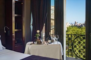 Esta habitación GL Monumento del Hotel Casa Fuster ofrece un balcón con vistas al paisaje urbano de Barcelona y a sus frondosos árboles. Disfrute de un vino y una rosa en su mesa mientras se deleita con la elegancia de este refugio. - ForTwo - Planes para parejas
