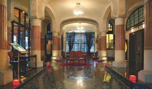 El elegante vestíbulo del Hotel Casa Fuster GL Monumento en Barcelona exhibe arcos ornamentados, pisos pulidos y sillas sobre una alfombra roja bajo una iluminación cálida, fusionando maravillosamente el encanto clásico con la comodidad moderna. - ForTwo - Planes para parejas