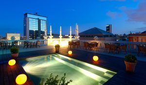 Al caer la noche en la terraza de la azotea del Hotel Casa Fuster GL Monumento (Barcelona), la piscina brilla junto a orbes luminosos y plantas en macetas, con mesas que ofrecen una vista relajante del cautivador horizonte de Barcelona. - ForTwo - Planes para parejas