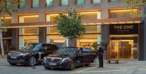 En el Hotel The One Barcelona GL, un aparcacoches abre con elegancia la puerta de un elegante coche negro en la entrada, mientras un opulento SUV está aparcado cerca. - ForTwo - Planes para parejas