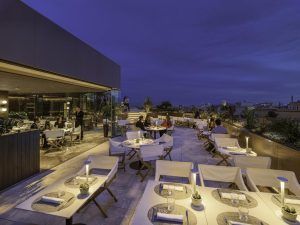 Al atardecer, el restaurante en la azotea del Hotel The One Barcelona GL ofrece una vista impresionante del paisaje urbano. Los comensales se relajan en mesas y sillas blancas, disfrutando del vibrante ambiente mientras el sol se pone sobre el encantador horizonte de Barcelona. - ForTwo - Planes para parejas