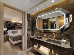 Este moderno baño, que evoca la lujosa suite GL del Hotel The One Barcelona, cuenta con un gran espejo octogonal y lavabos dobles. Desde aquí, se vislumbra el dormitorio, con una elegante pared con detalles florales que le da un toque de estilo ejecutivo. - ForTwo - Planes para parejas