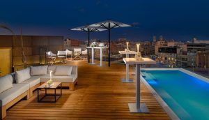 El patio de la azotea del Hotel The One Barcelona GL cuenta con asientos, mesas, sombrillas y una piscina resplandeciente que complementa maravillosamente el horizonte de la ciudad por la noche. - ForTwo - Planes para parejas
